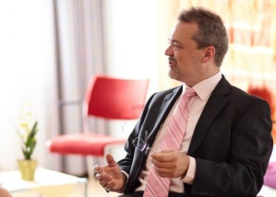 Professor Dr. Bielitz sitzt mit Brille in der Hand auf einem Stuhl und spricht.